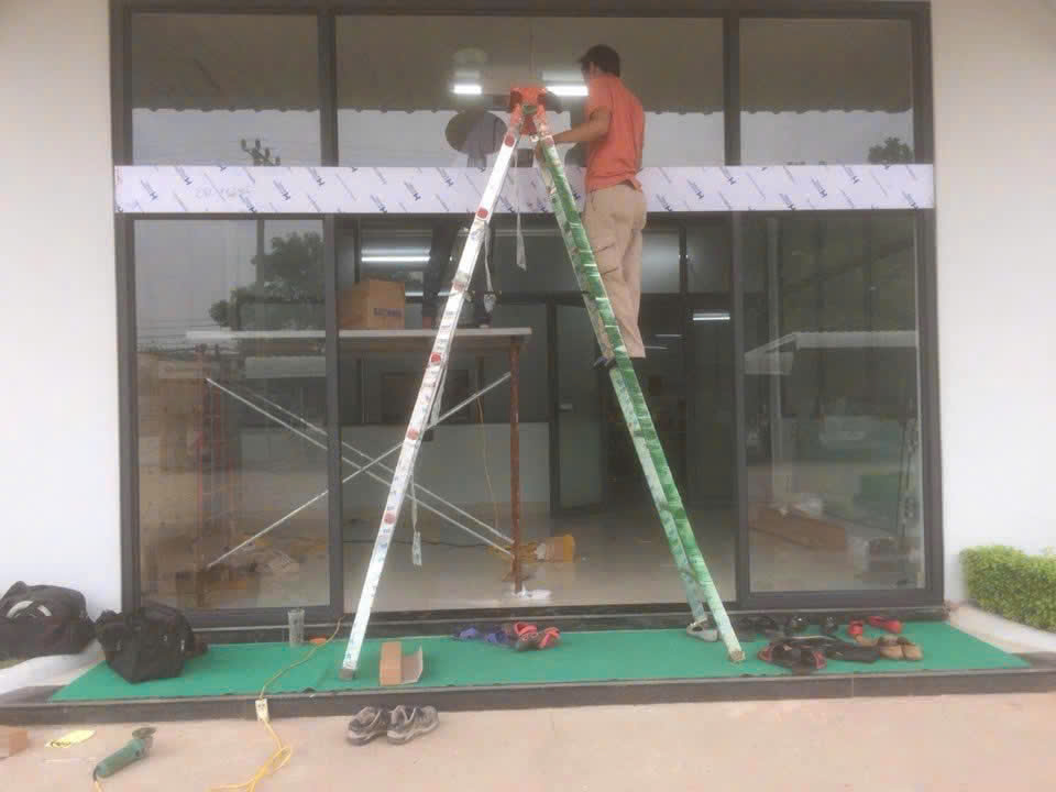 Cửa kính cường lực mặt tiền đang được thợ lắp đặt bằng thang và dụng cụ thi công